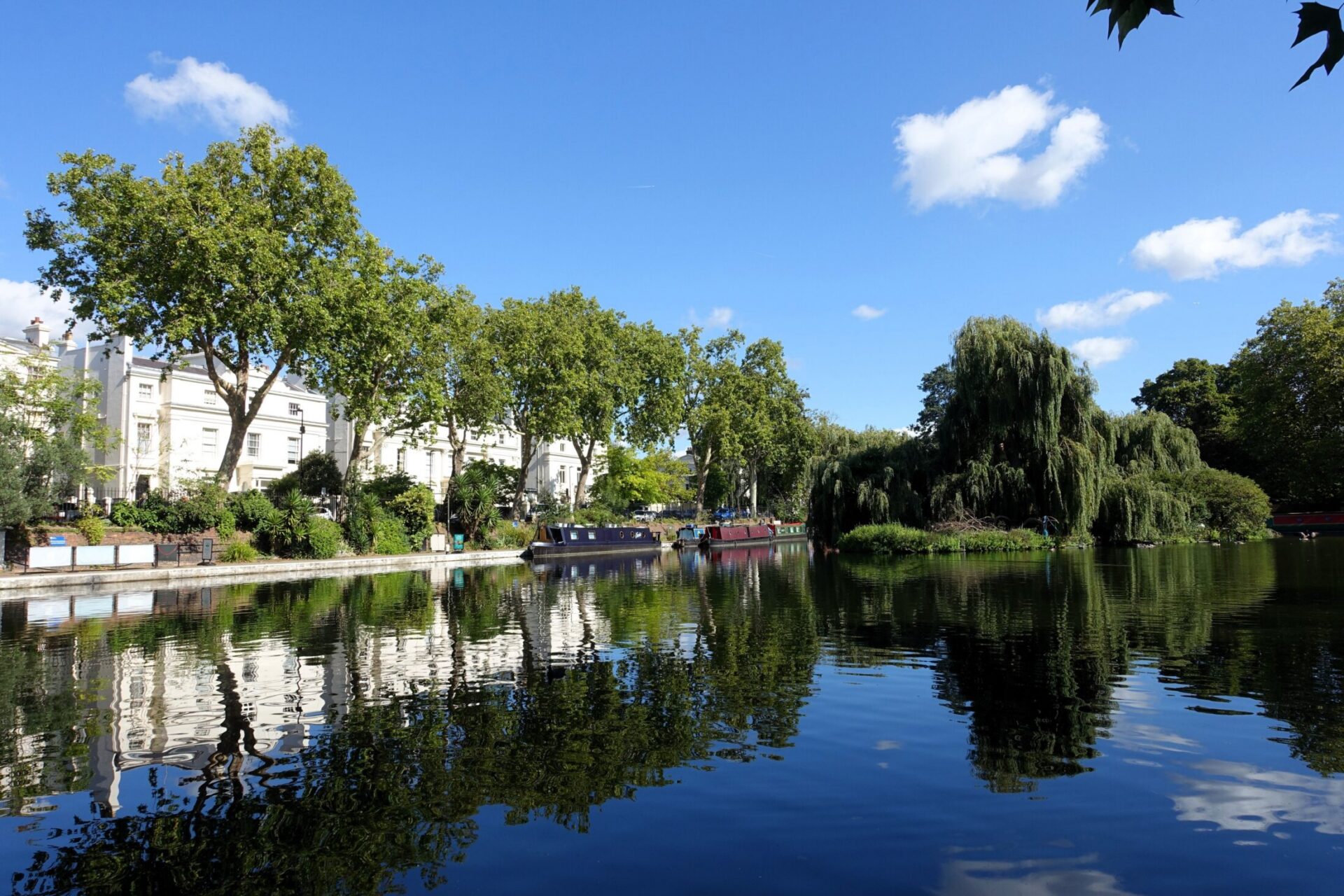 Tranquil canal in Little Venice, London with boats, trees, and reflections under a clear blue sky.